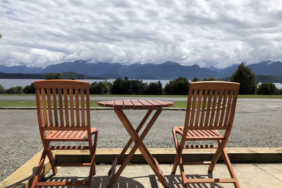 Two wooden chairs and a small table on a gravel surface with a view of mountains and a lake.
