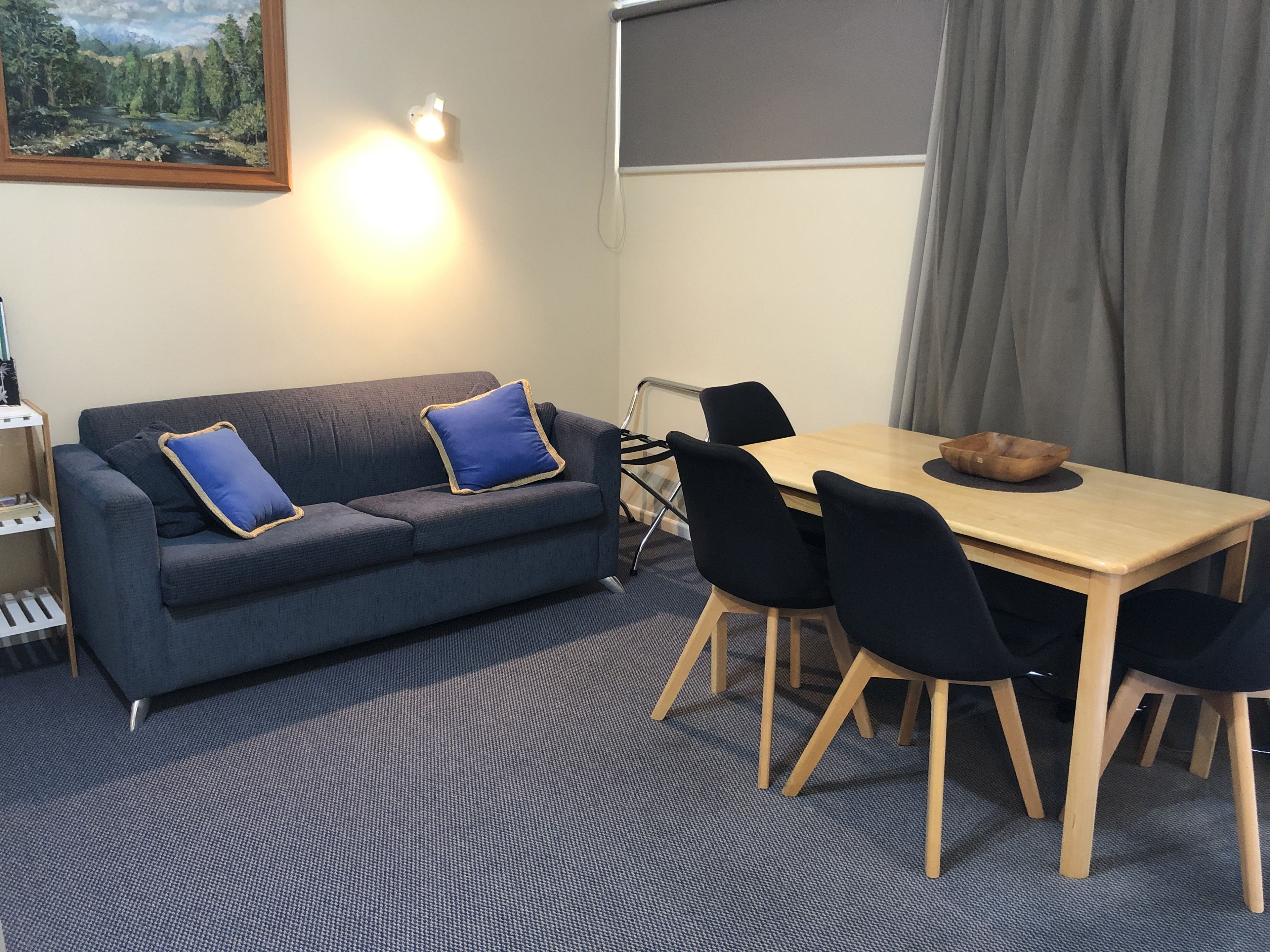 Quarto com um sofá azul escuro, duas almofadas azuis, uma mesa de jantar de madeira com quatro cadeiras e um quadro na parede.