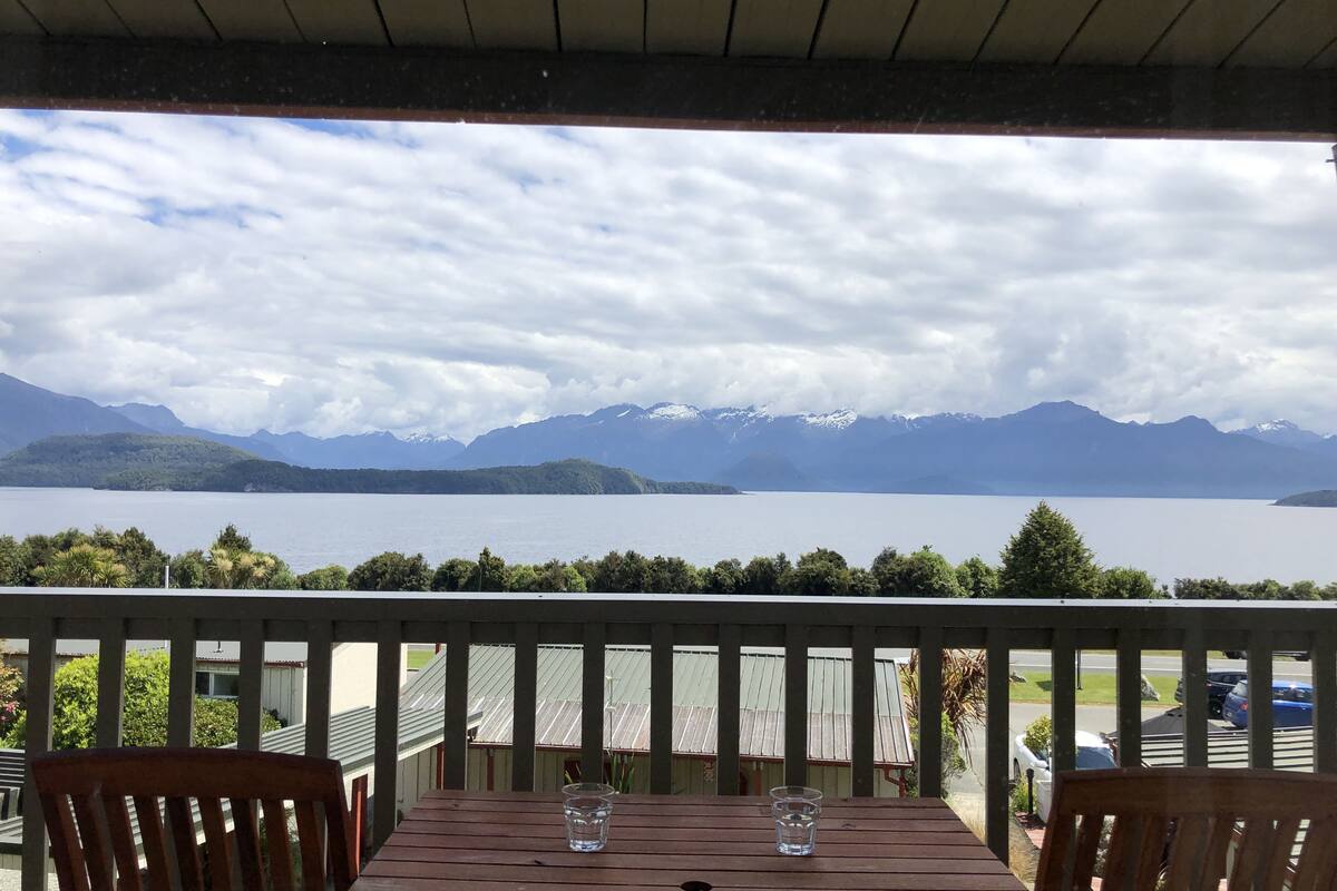 A wooden table with two glasses on a balcony overlooking a lake and mountains.