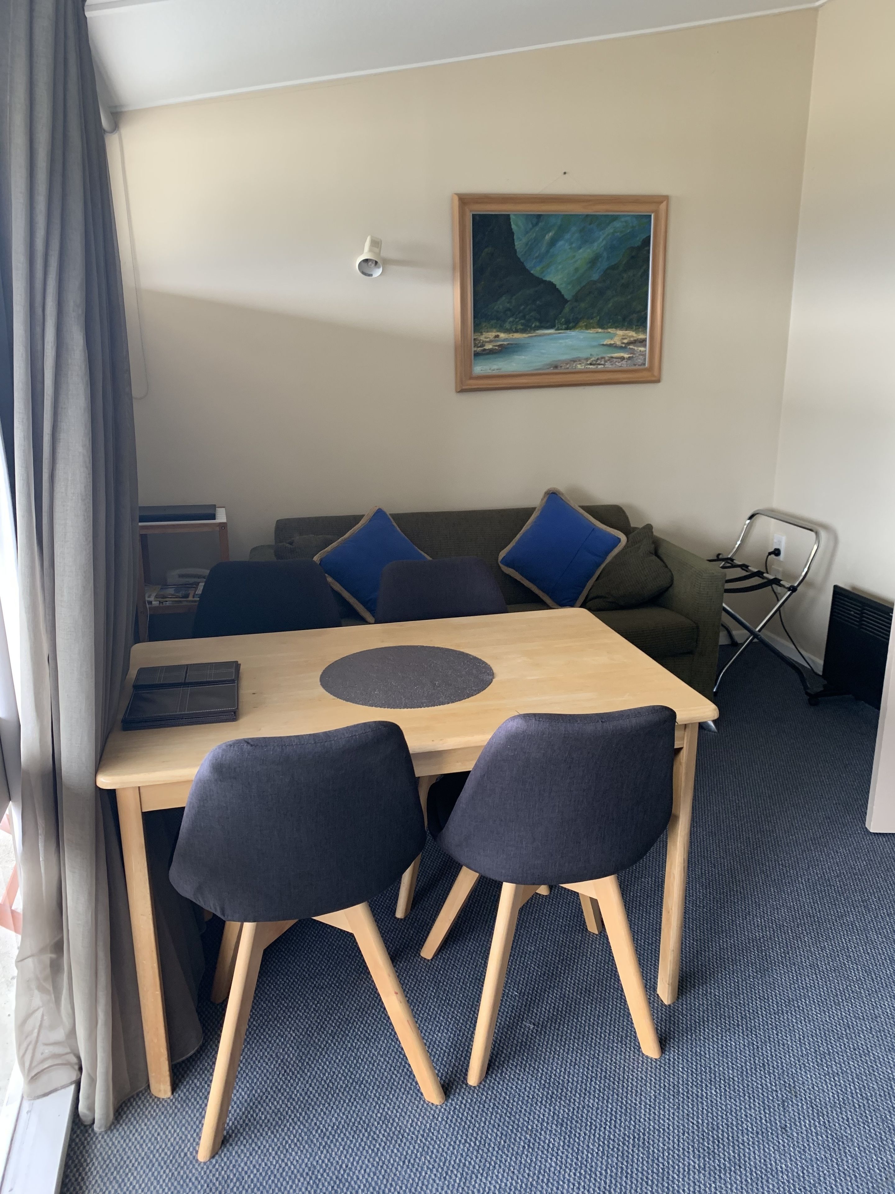 A room with a wooden table, two chairs, a sofa with blue cushions, a painting on the wall, and a window with curtains.