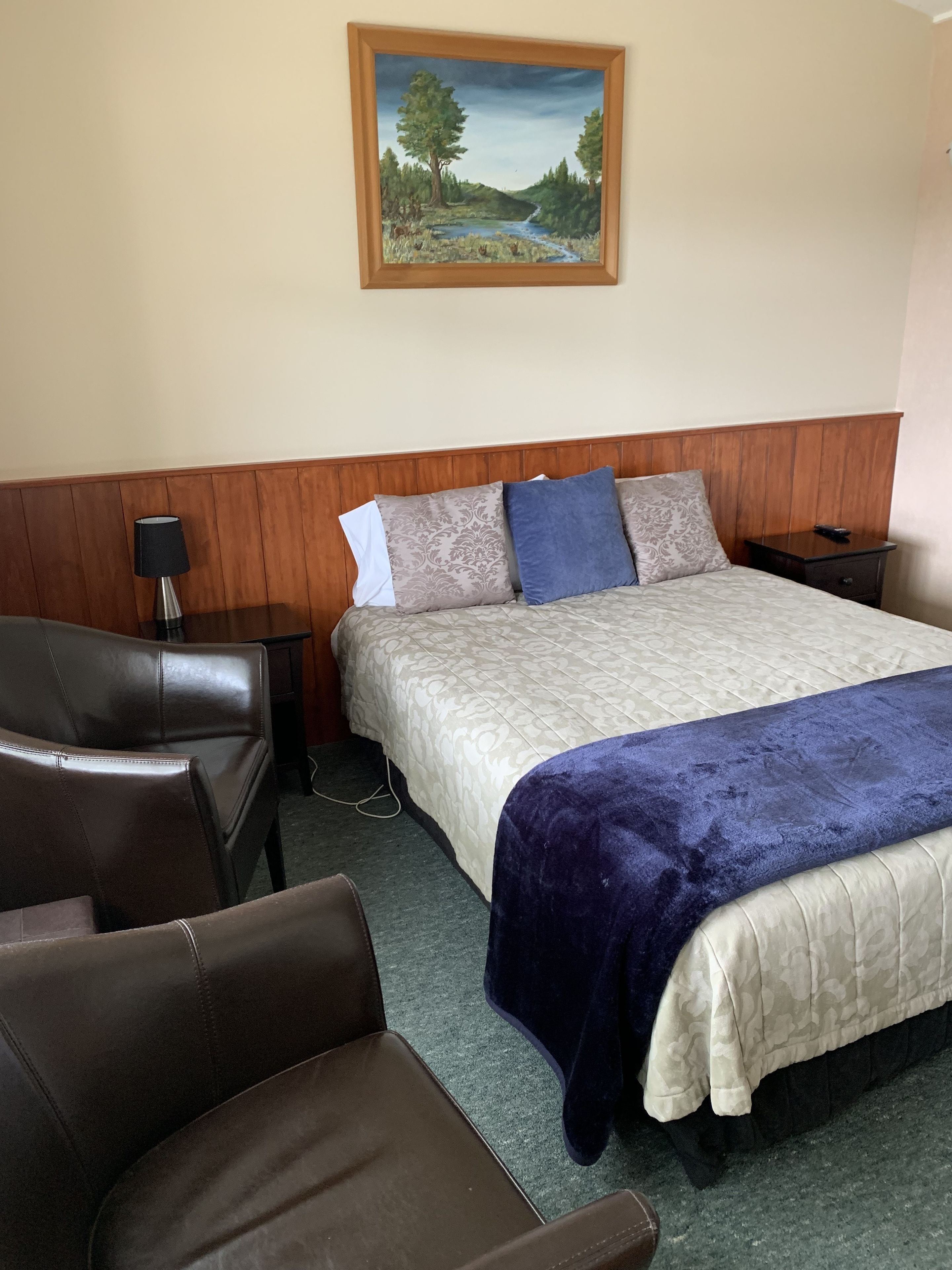A hotel room with a bed, a wooden headboard, a nightstand, a lamp, a chair, and a painting of a landscape on the wall.