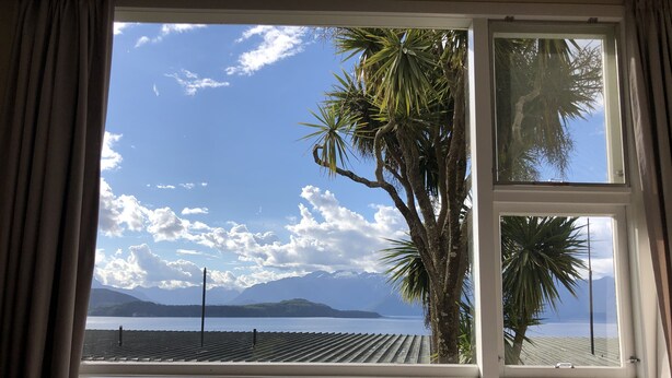 A room with a large window overlooking a body of water, palm trees, and distant mountains. - Te Anau Manapouri Lake Hotel (Manapouri)