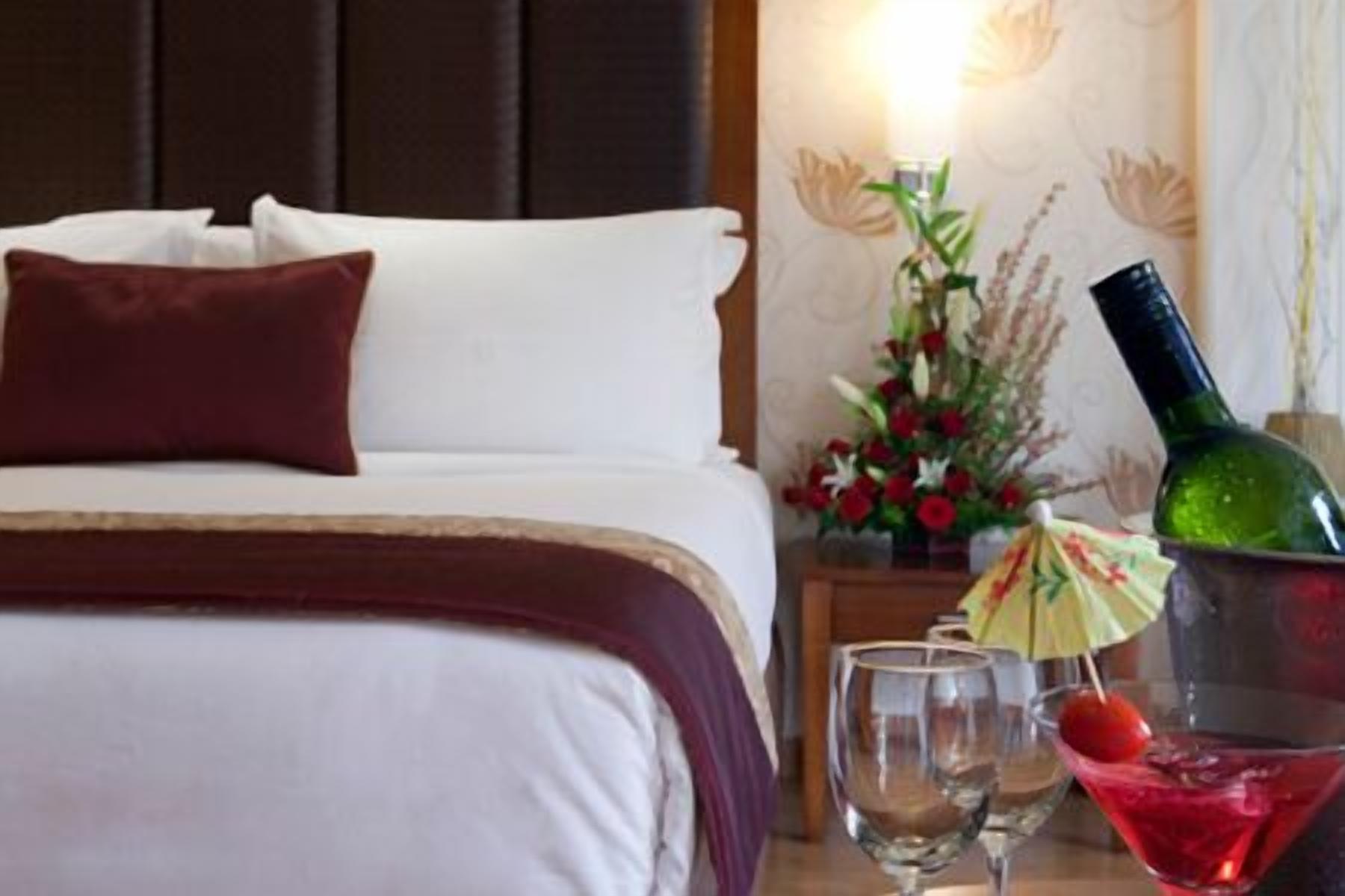 Quarto de hotel com cama, almofadas, mesa de cabeceira com um vaso de flores, uma garrafa e dois copos.
