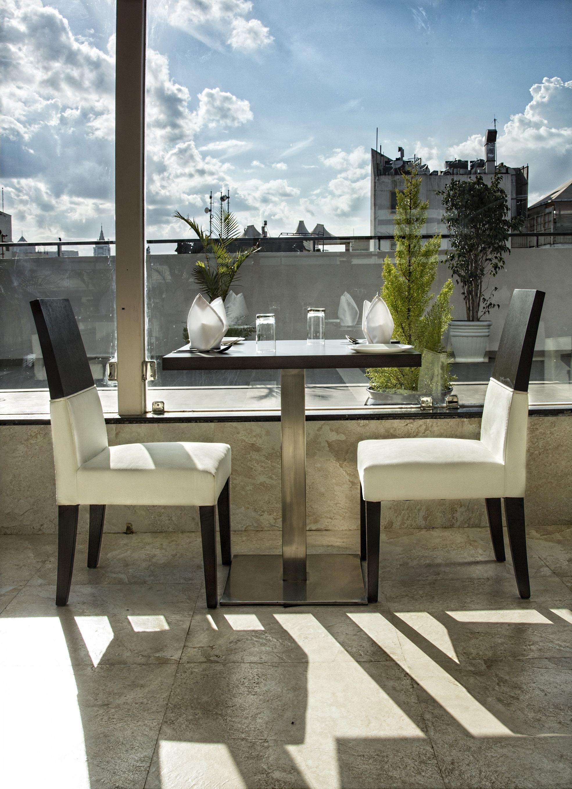 A rooftop terrace with a table and two chairs, potted plants, and a view of the cityscape.