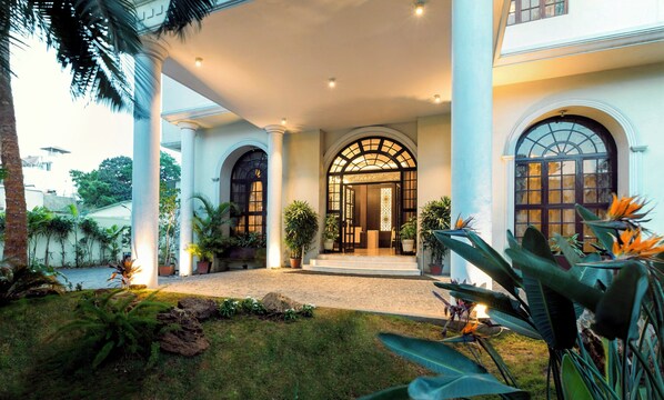 Uma entrada imponente com galeria coberta, grandes janelas em arco e exuberante vegetação tropical.