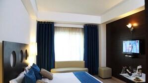 A modern hotel room with a large bed, a TV, a small table with a bottle and glasses, and a window with curtains. - Silver Ferns (New Delhi)