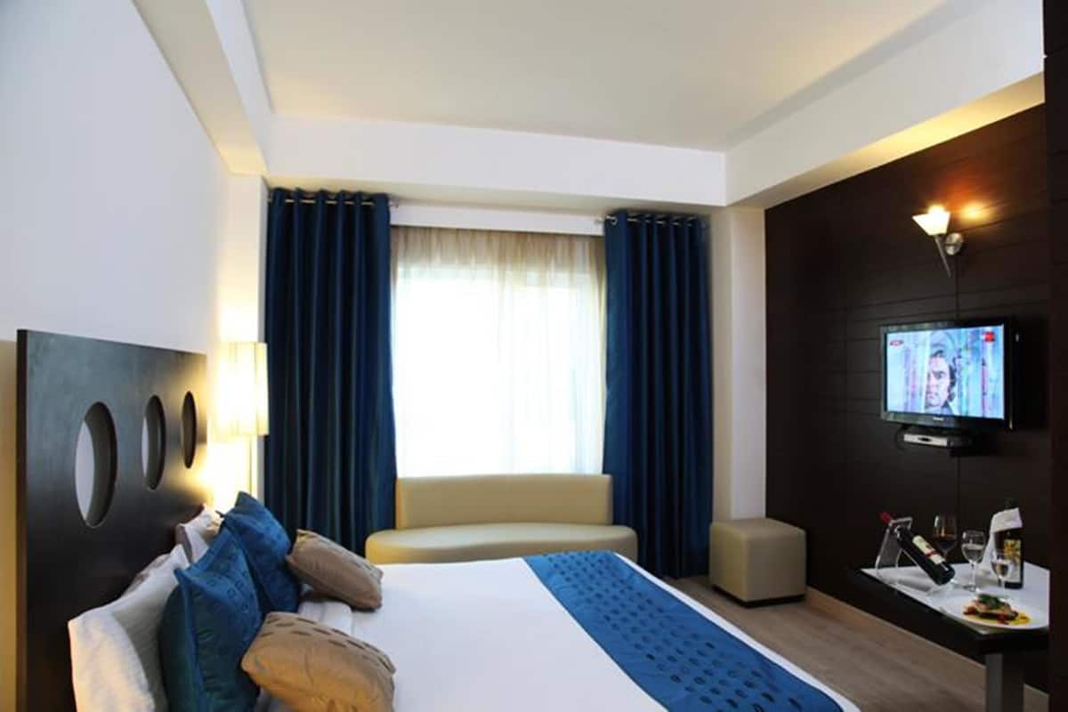 A modern hotel room with a large bed, a TV, a small table with a bottle and glasses, and a window with curtains.