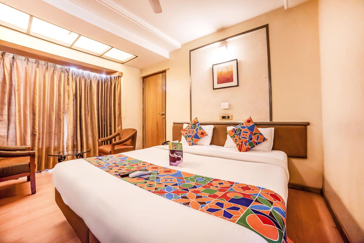 Hotelkamer met een groot bed, een bureau en een stoel. Er is een raam met gordijnen, een schilderij aan de muur en een plafondventilator.