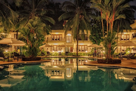 A pool area with lounge chairs and umbrellas, surrounded by palm trees and a building with multiple balconies.