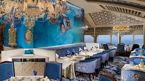 A luxurious dining area with ornate chandeliers, blue upholstered chairs, and a high ceiling with intricate patterns. - Golkonda Resorts & Spa (Hyderabad)