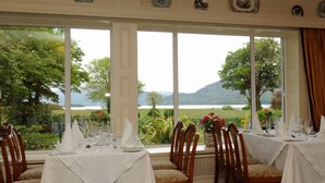 A dining area with tables set for a meal, large windows offering a view of a lake and greenery. - Loch Lein Country House (Killarney)