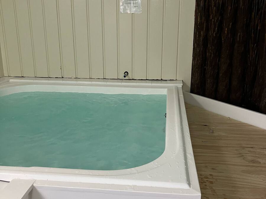 Uma banheira de hidromassagem retangular com exterior branco, rodeada por um deck de madeira e uma parede branca com painéis verticais.