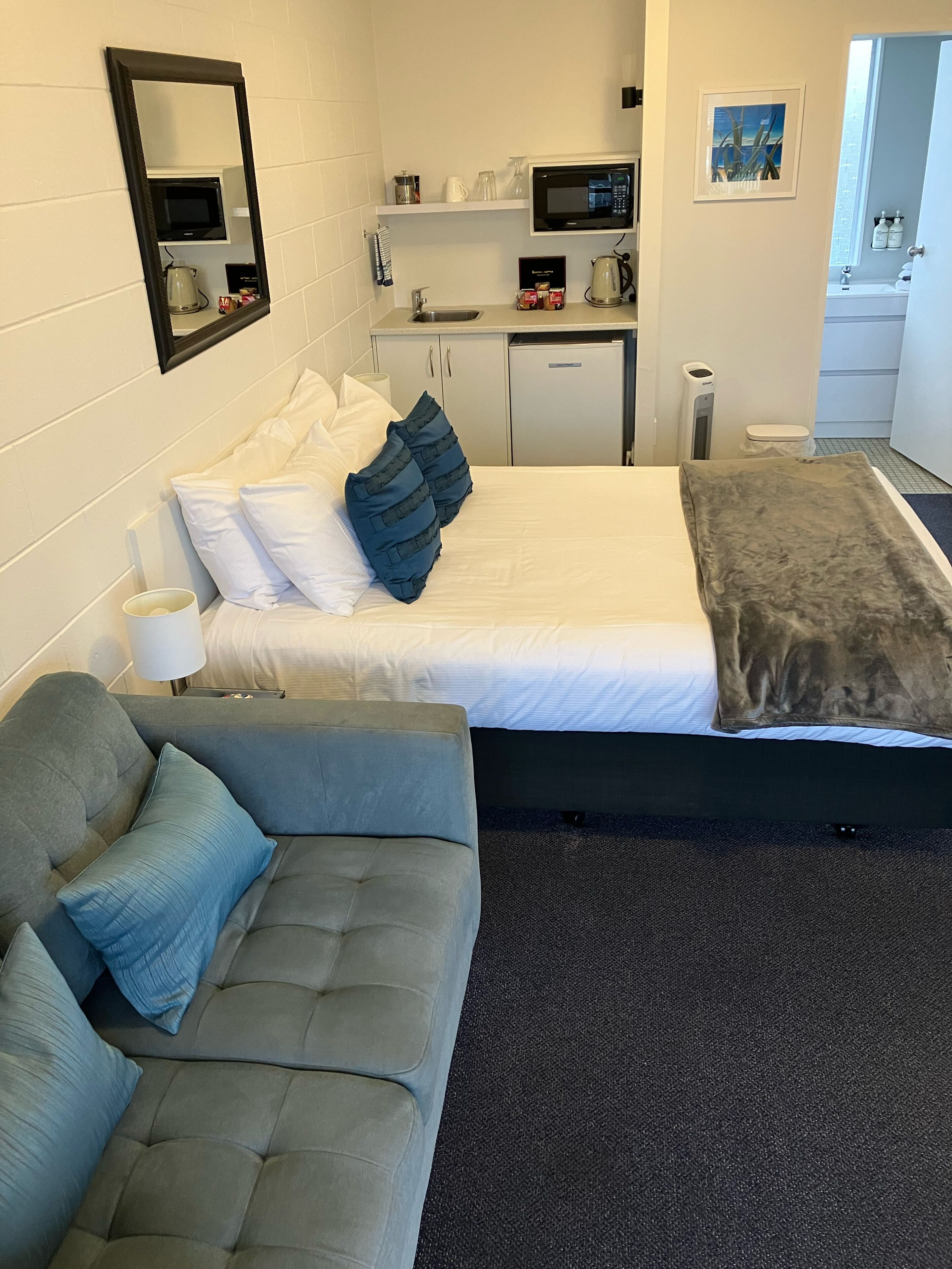 Ein Hotelzimmer mit Bett, einer Sitzecke, einer kleinen Küchenzeile und einem durchgehenden Tür sichtbar liegenden Badezimmer.