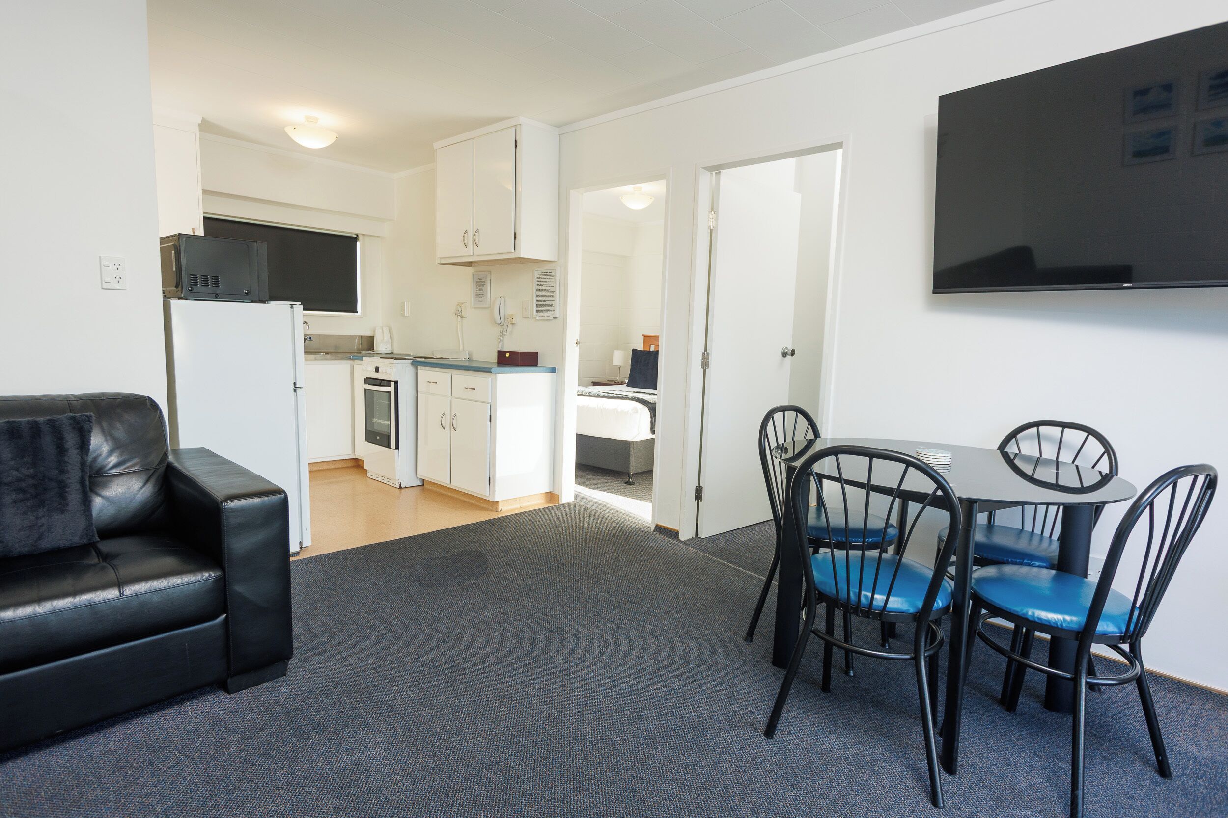Un espace de vie compact comprenant un coin cuisine, un espace repas avec une table et des chaises, un canapé en cuir noir et un téléviseur à écran plat fixé au mur.
