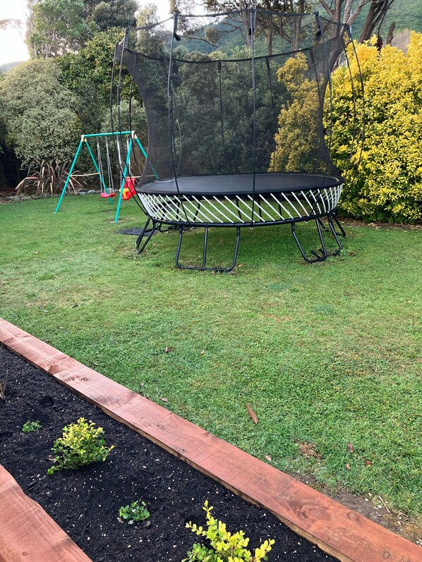 Un trampolín rodeado por una red de seguridad, un conjunto de columpios y una valla de madera con plantas.