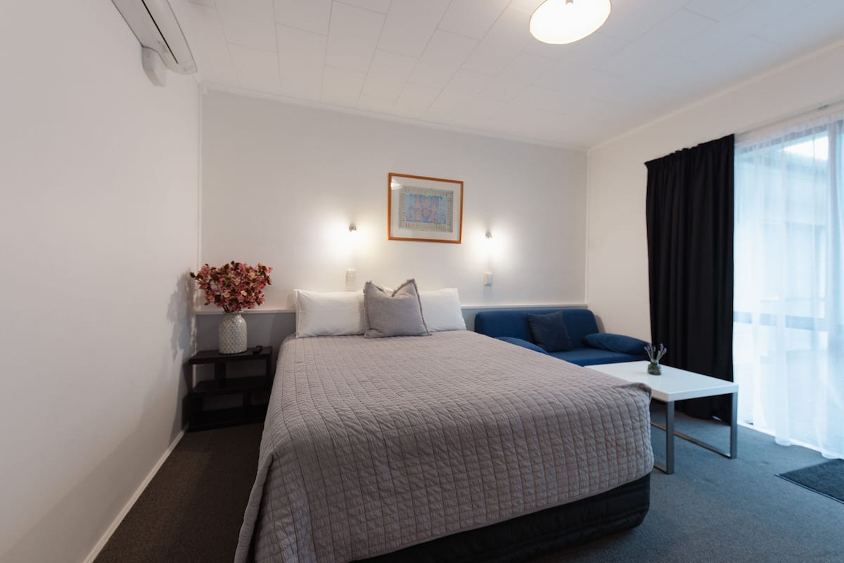 A modern hotel room with a large bed, a small table, a sofa, and a window with curtains.