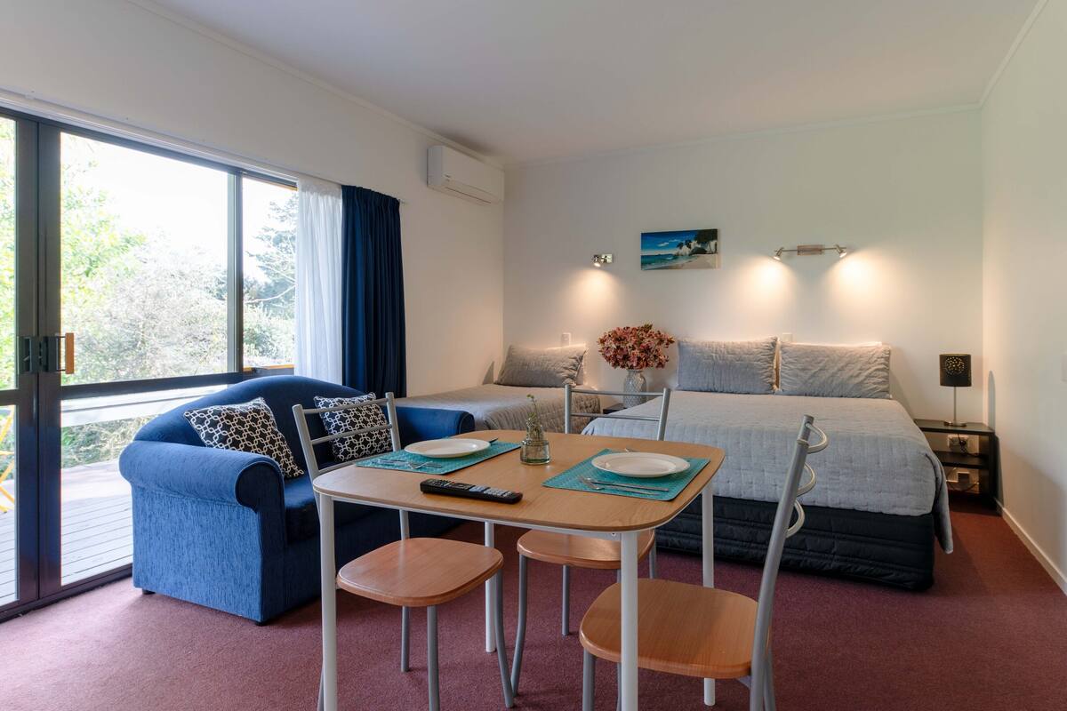 A modern hotel room with a large bed, a blue sofa, a dining table with chairs, and a view through the window.