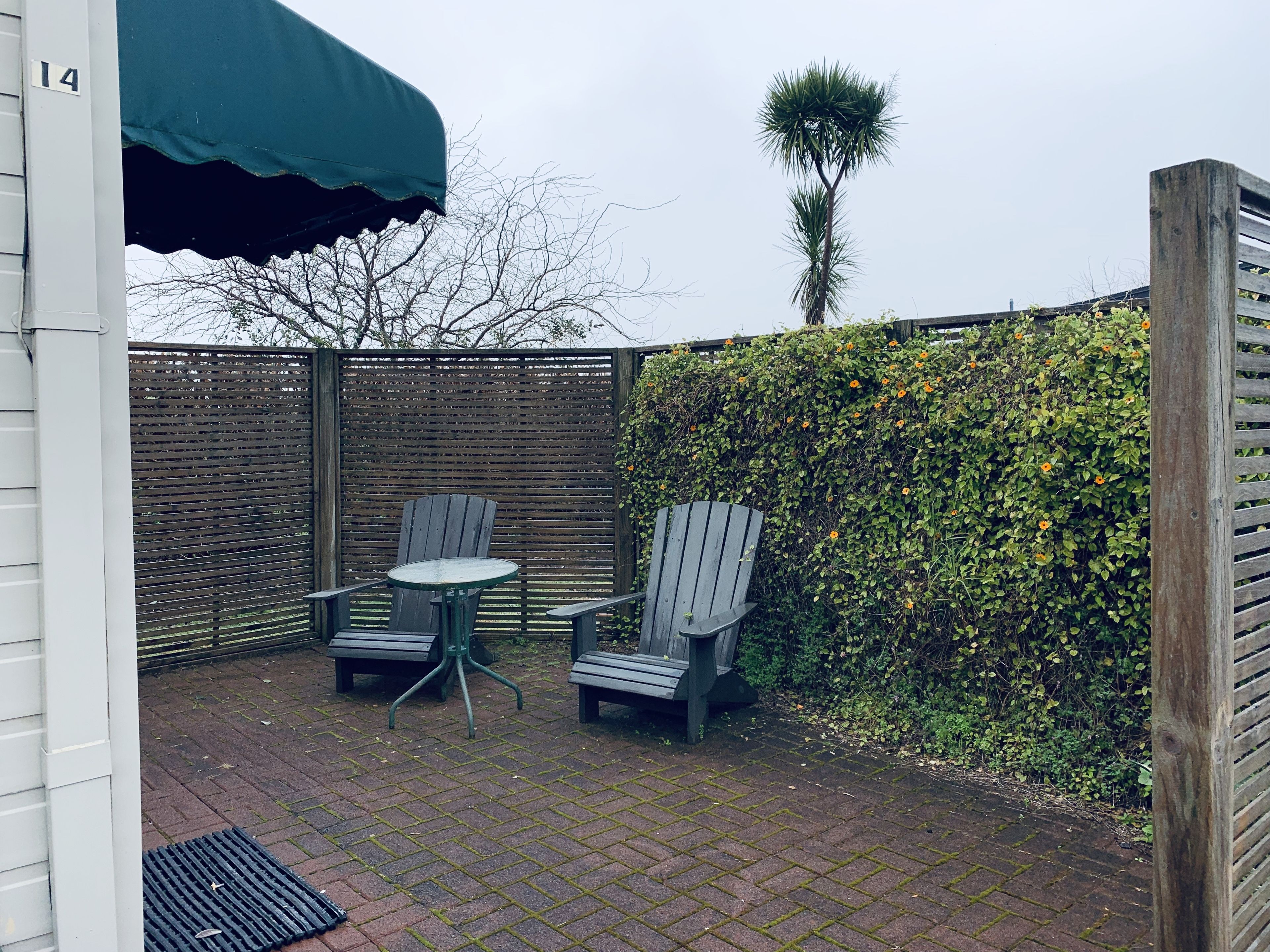 A patio with two wooden chairs and a small table, a green umbrella, and a hedge with orange flowers.
