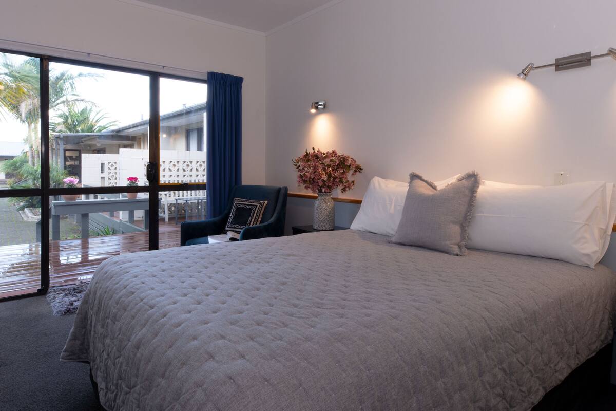 A bedroom with a large bed, a chair, a vase with flowers, and a view of the outdoors.