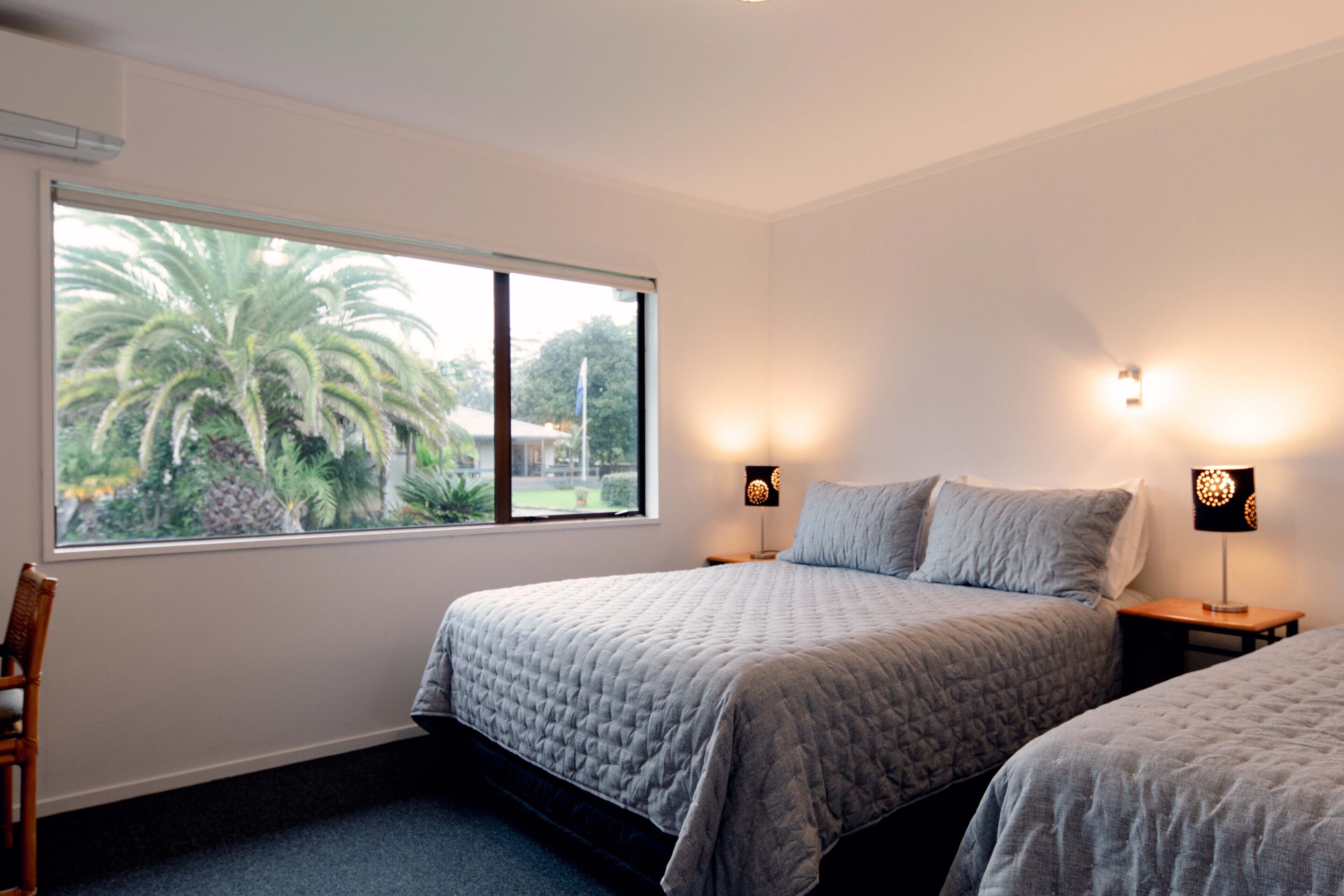 A bedroom with two beds, a window with a view of palm trees, and two bedside lamps.