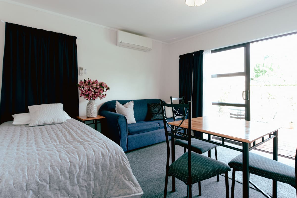 A hotel room with a bed, a sofa, a dining table with chairs, a vase with flowers, and a sliding glass door to the outside.