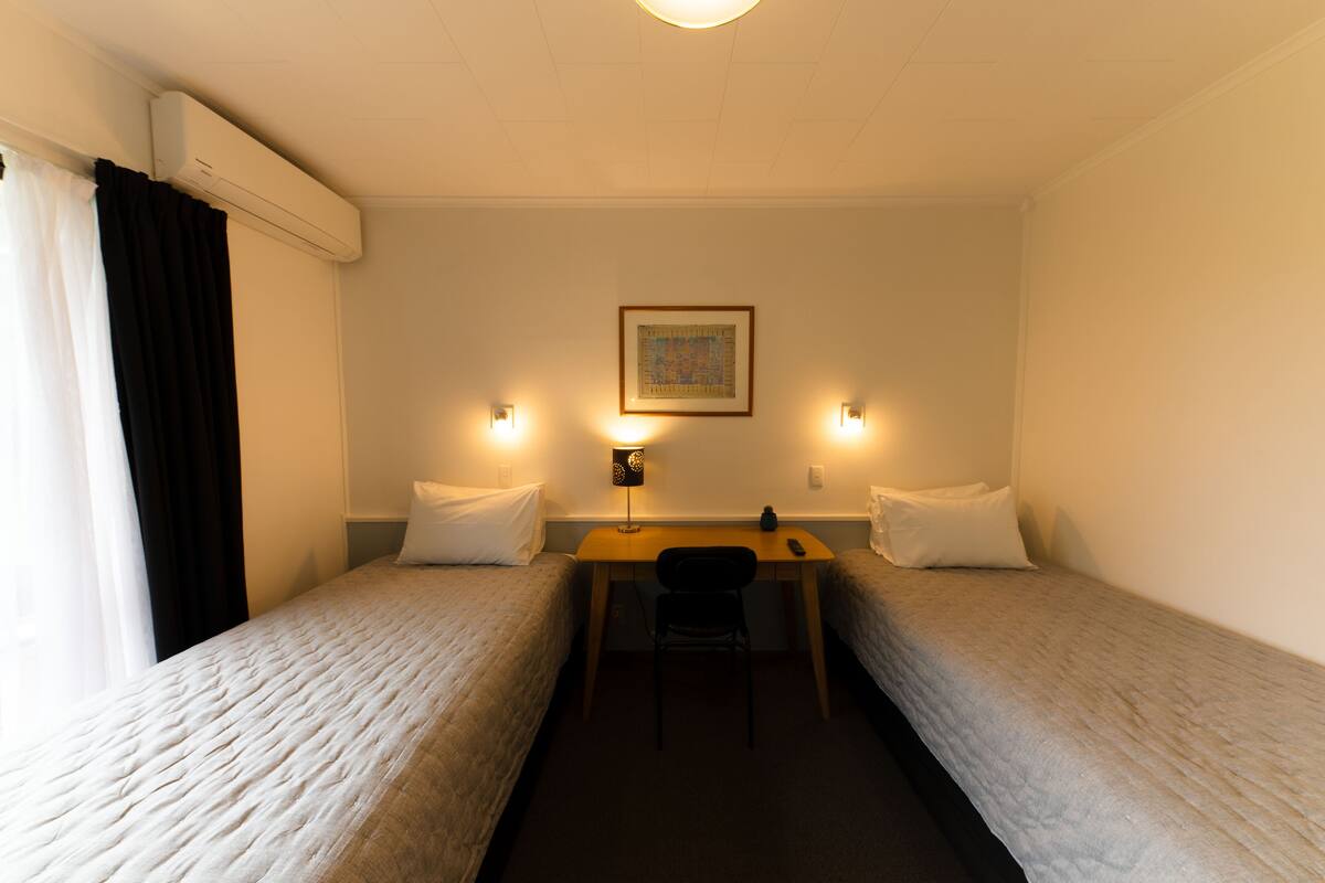 A hotel room with two single beds, a desk, a chair, and a wall-mounted air conditioner.