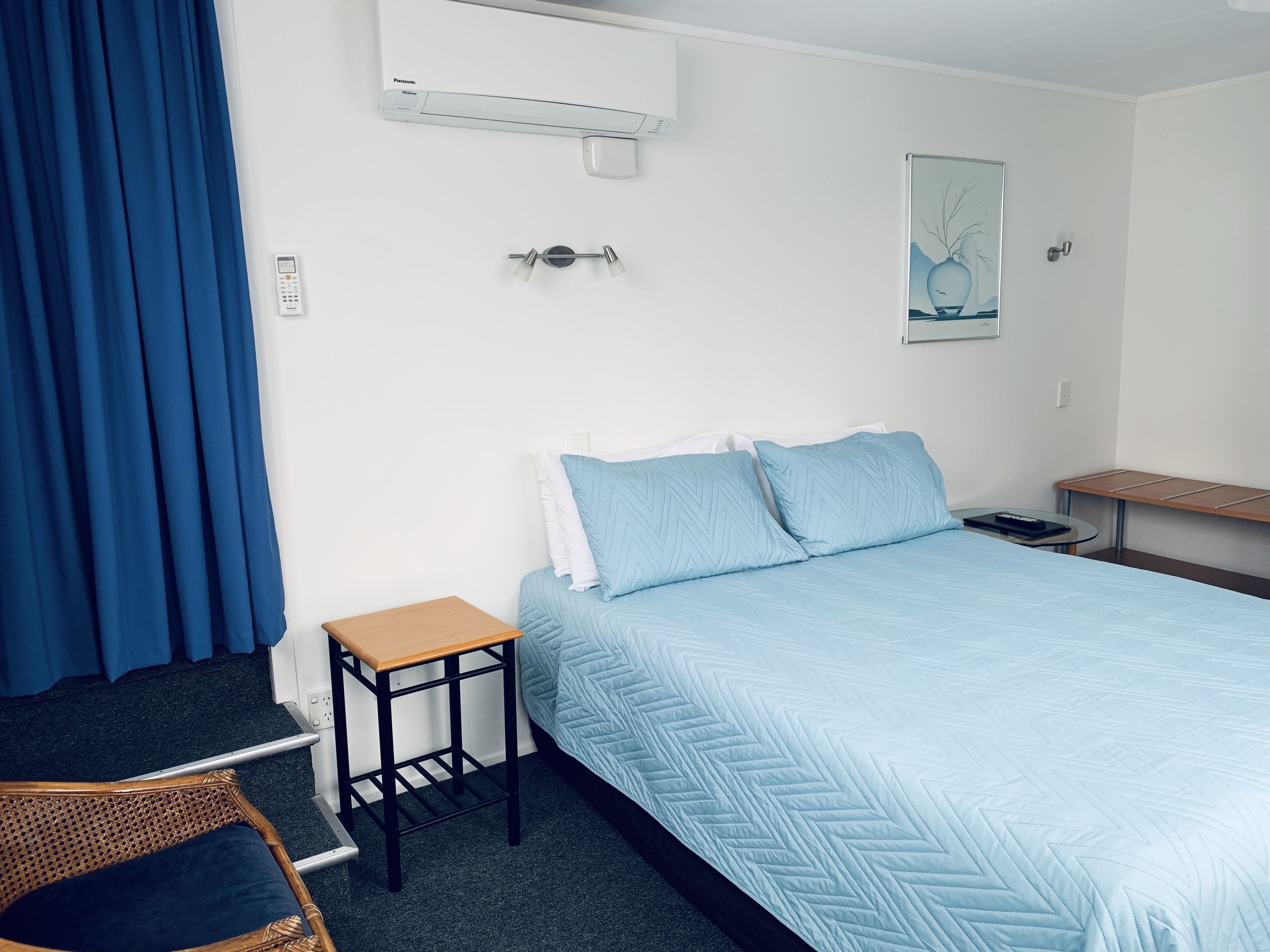 A hotel room with a bed, a small table, a chair, an air conditioning unit, a power outlet, and a painting on the wall.