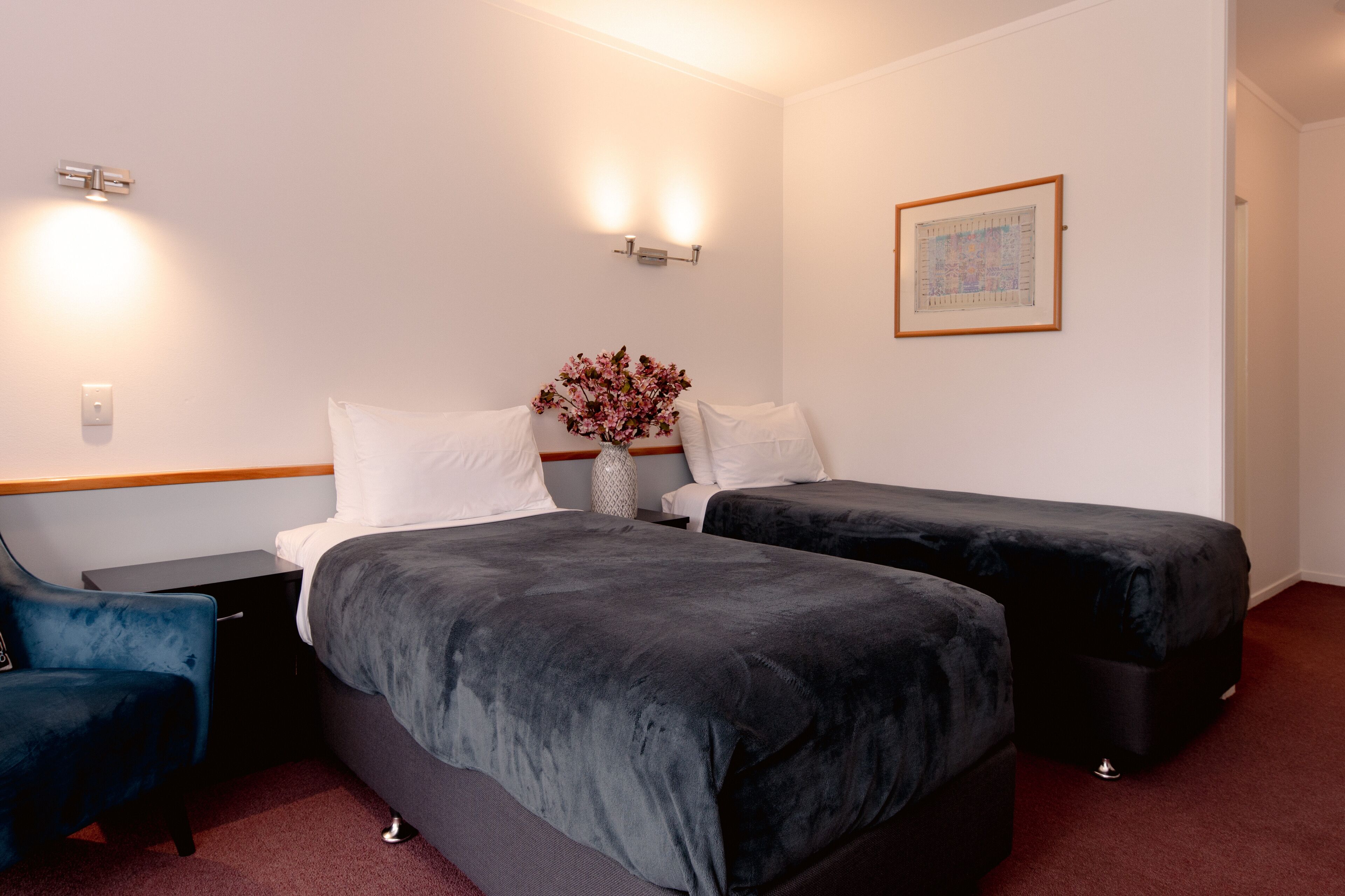 A hotel room with two beds, a nightstand, a chair, and a vase with flowers.