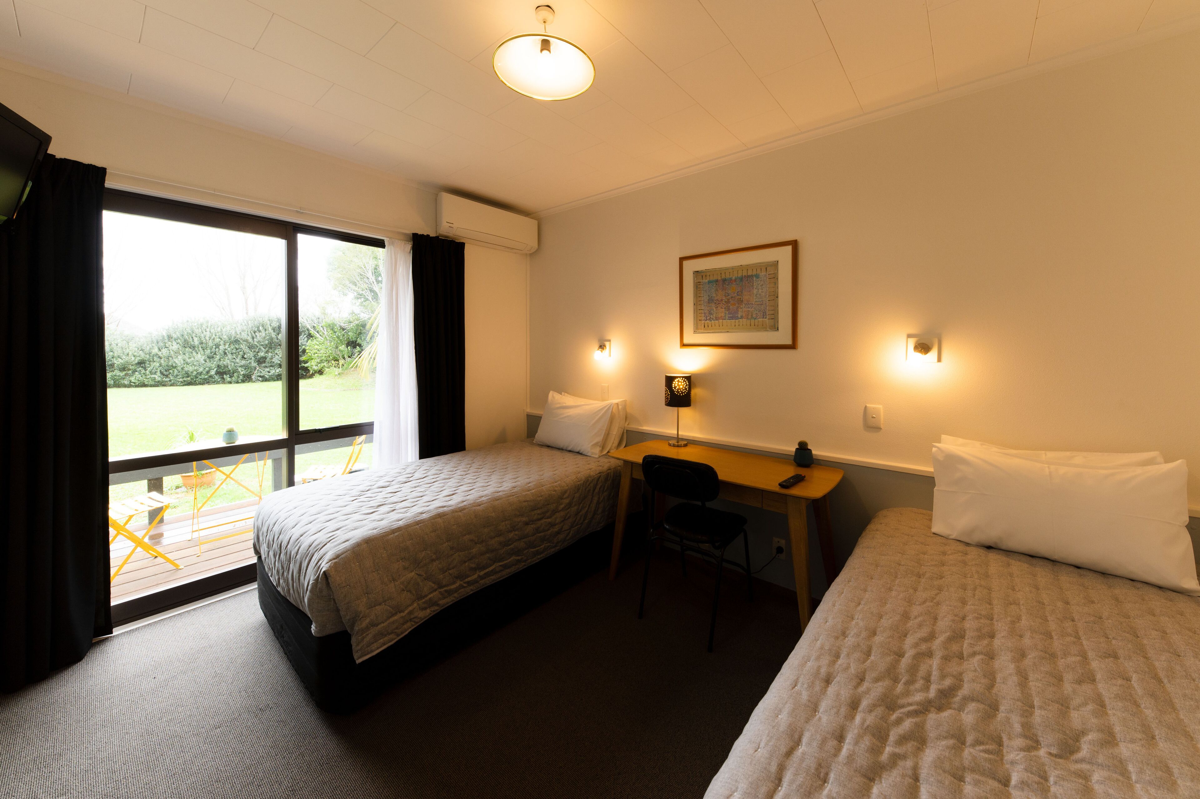 A hotel room with two beds, a desk, a chair, a window with curtains, a wall-mounted light, and a framed picture.
