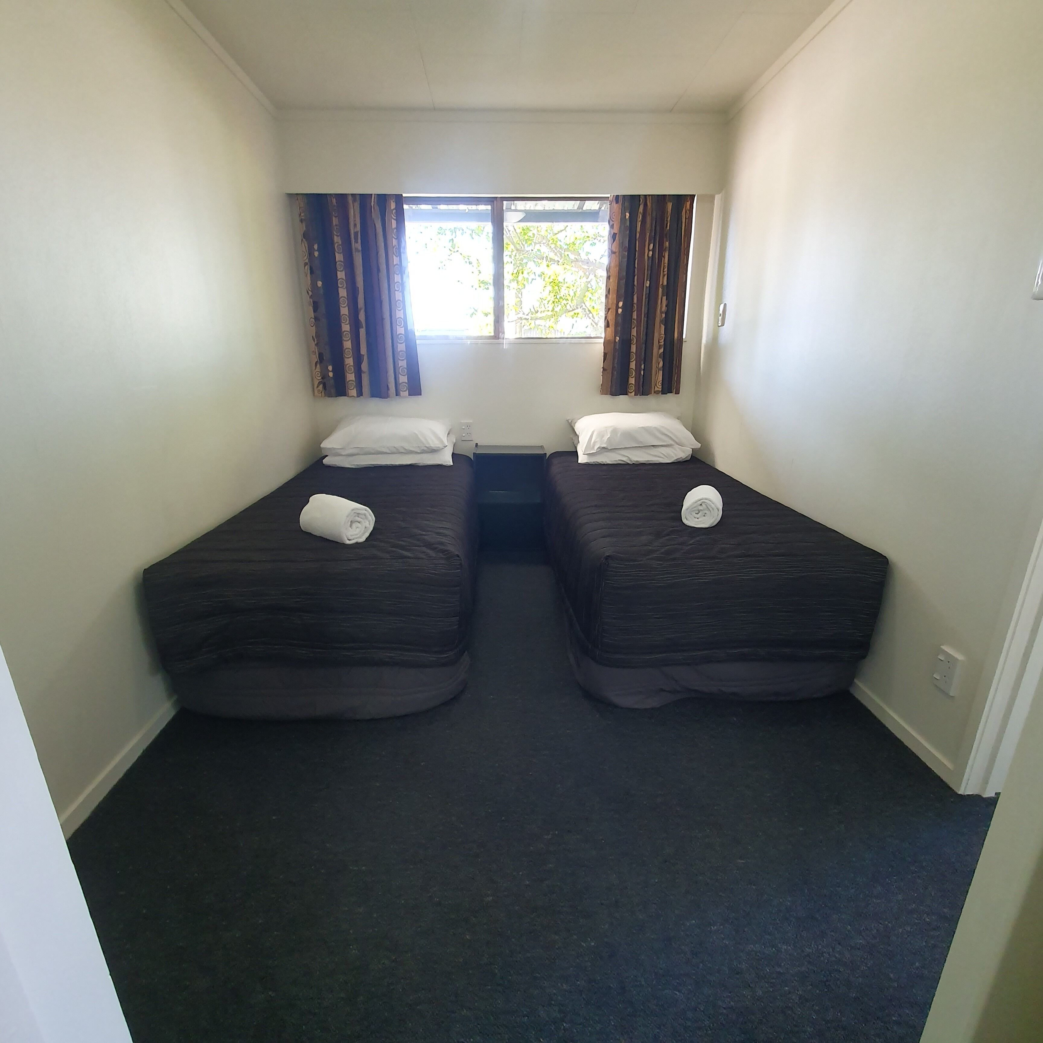 Une petite chambre propre, avec deux lits simples, une fenêtre avec des rideaux et un sol recouvert de moquette.