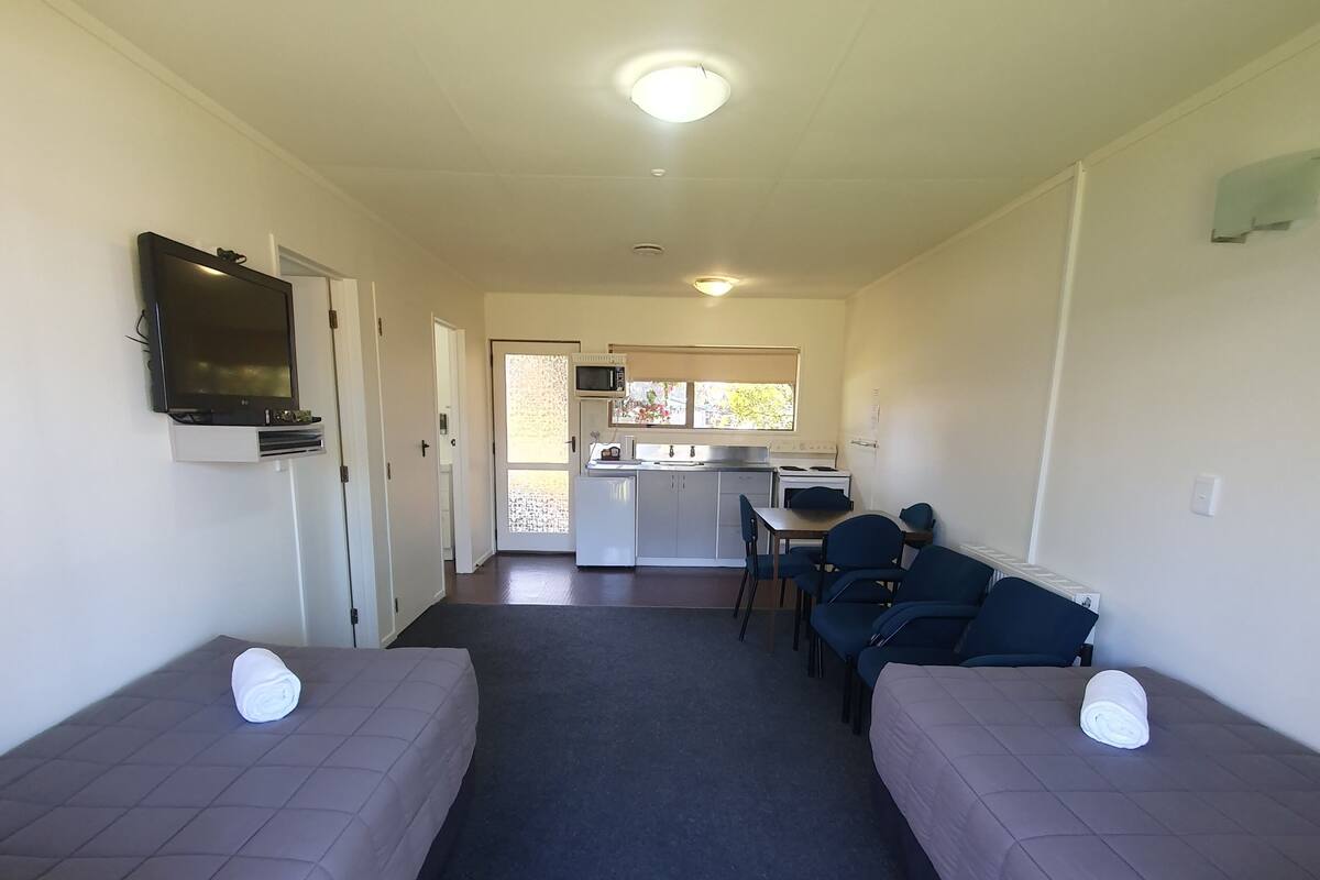 A compact hotel room with a kitchenette, a TV mounted on the wall, and two beds with pillows.