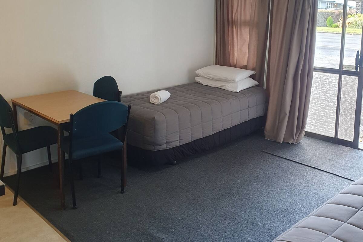 A small, well-lit room with a bed, a desk, and a chair. There is a window with curtains, and a ceiling light fixture.