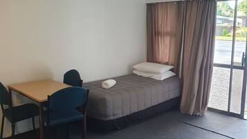 A small, well-lit room with a bed, a desk, and a chair. There is a window with curtains, and a ceiling light fixture.