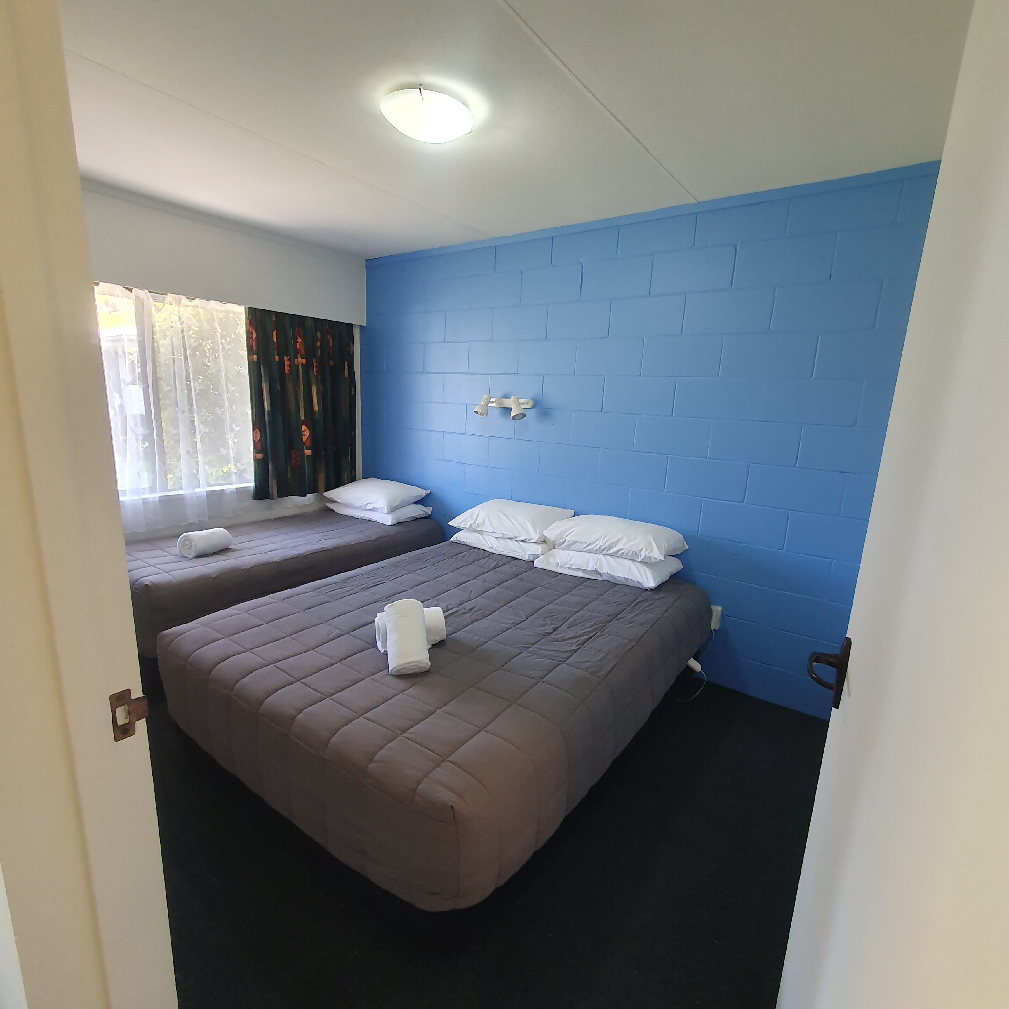 A hotel room with two beds, each with a brown quilted bedspread and white pillows. There is a window with sheer curtains and a decorative curtain partially drawn. The room has a blue brick-patterned wall and a white ceiling with a recessed light fixture.