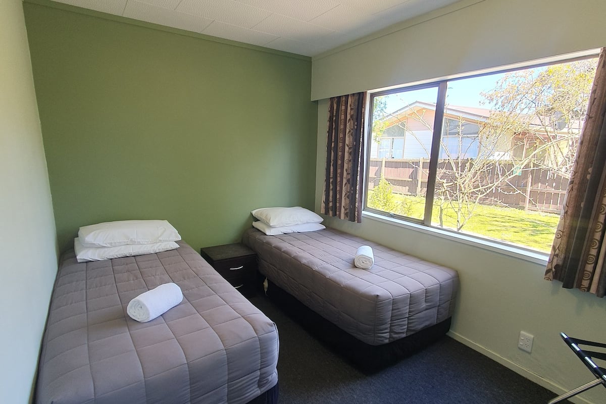 A small, clean room with two beds, a window with curtains, and a view of a house and garden outside.