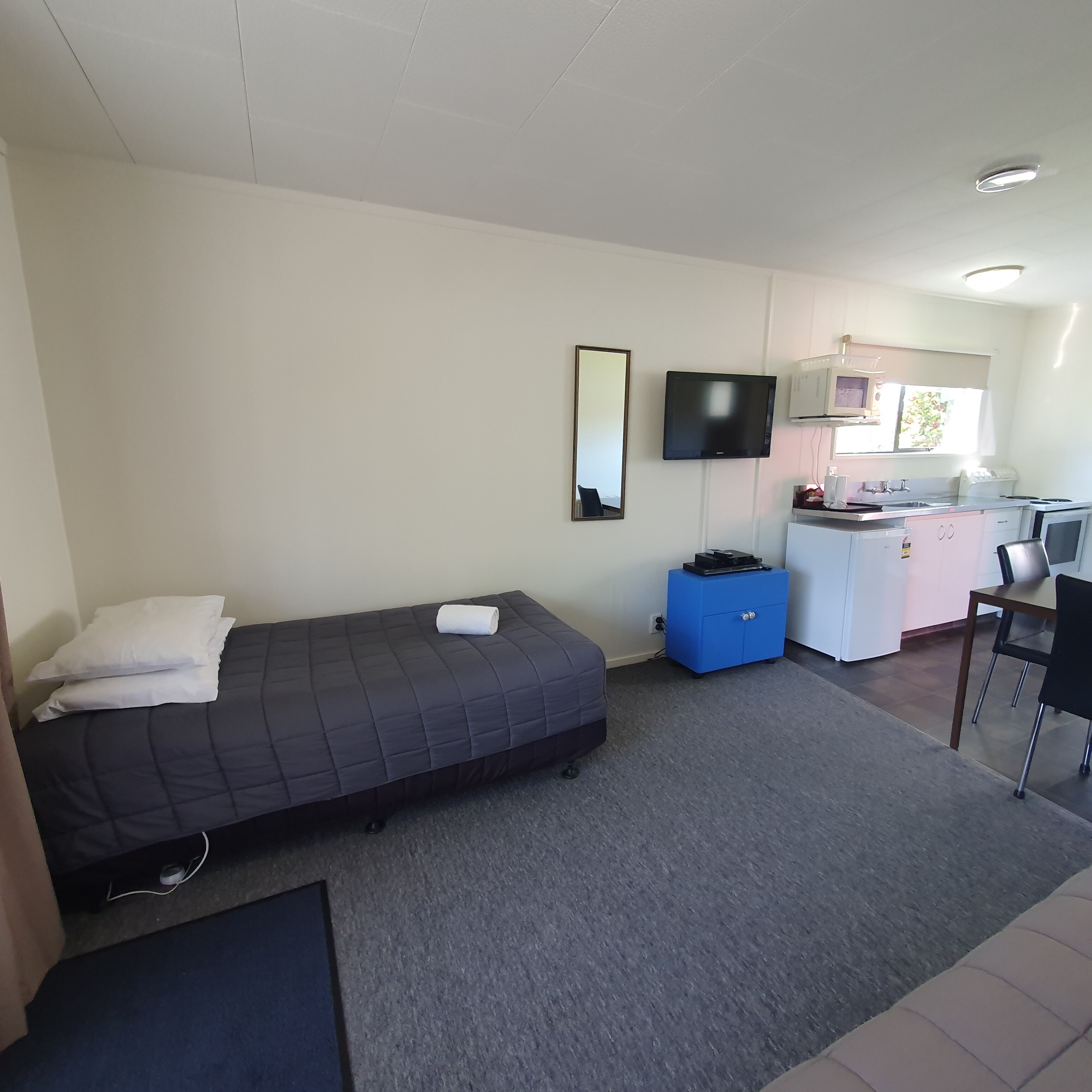 A compact room with a bed, a small desk, a chair, a TV, and a kitchenette.
