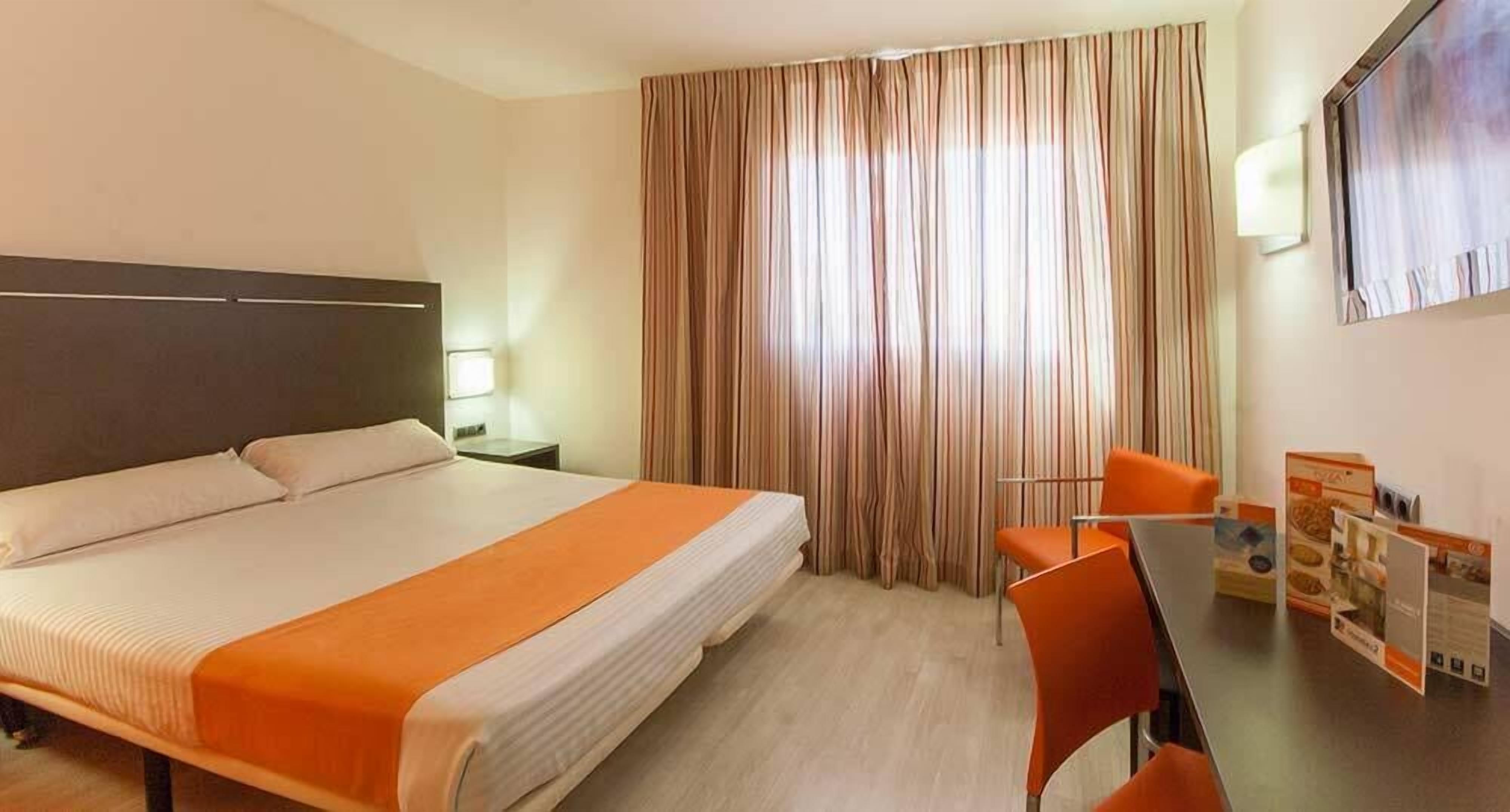 Habitació d'hotel amb un llit gran, una escriptori amb dues cadires, una televisió i cortines.