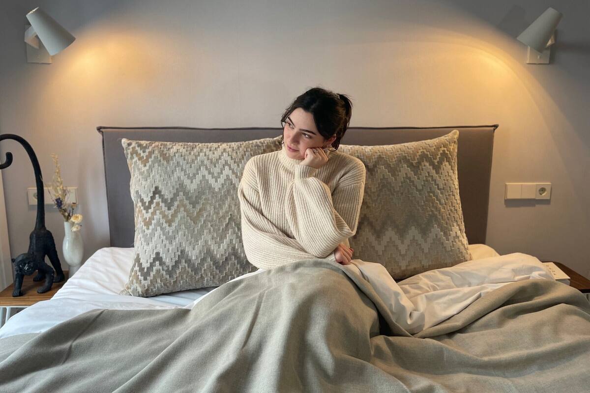 A person sitting on a bed with a patterned pillow, a blanket, and a wall-mounted light fixture.