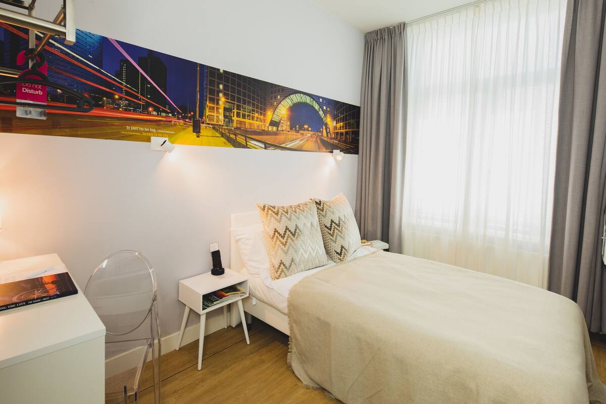 A hotel room with a bed, a desk, a chair, a nightstand, and a large cityscape painting on the wall.