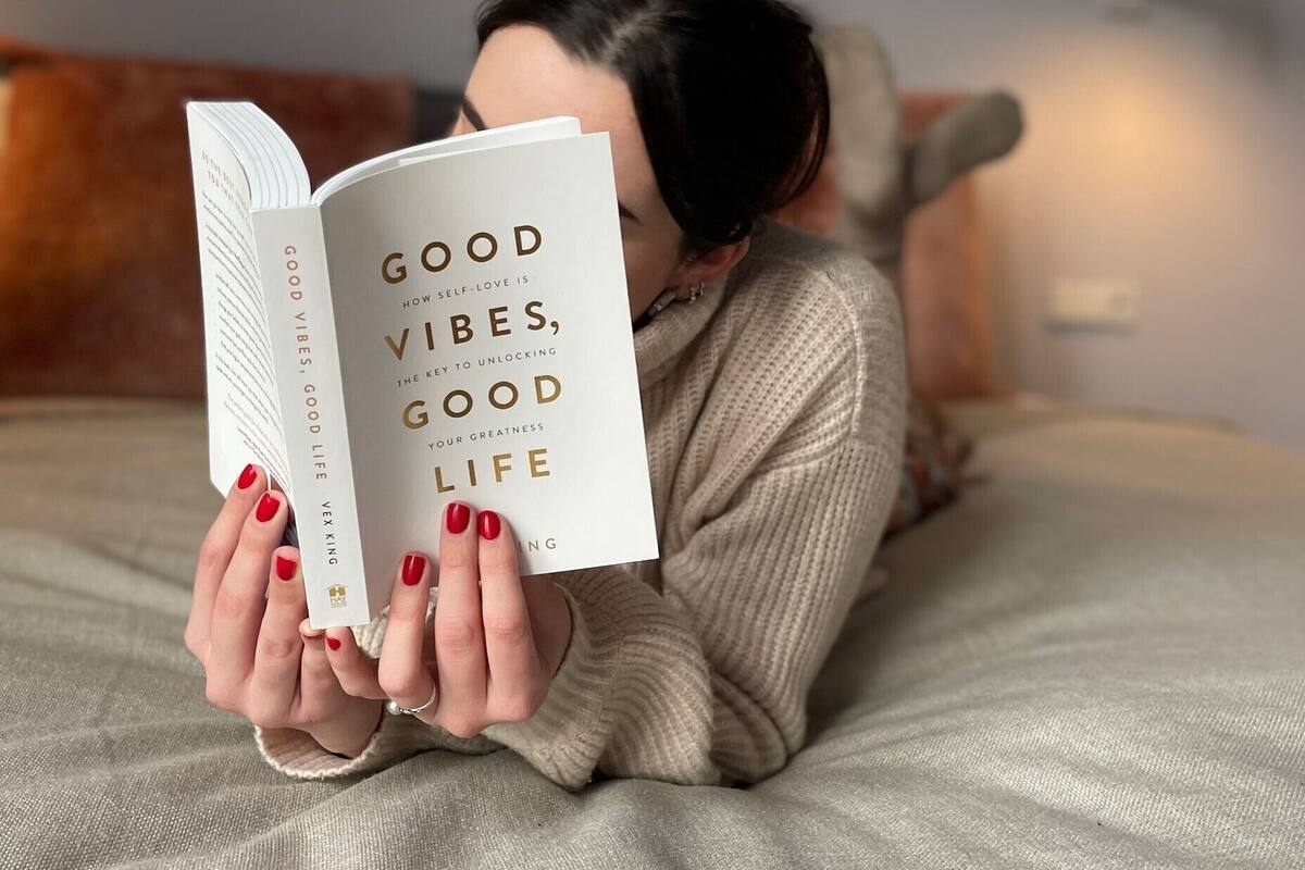A person lying on a bed reading a book titled "Good Vibes, Good Life".