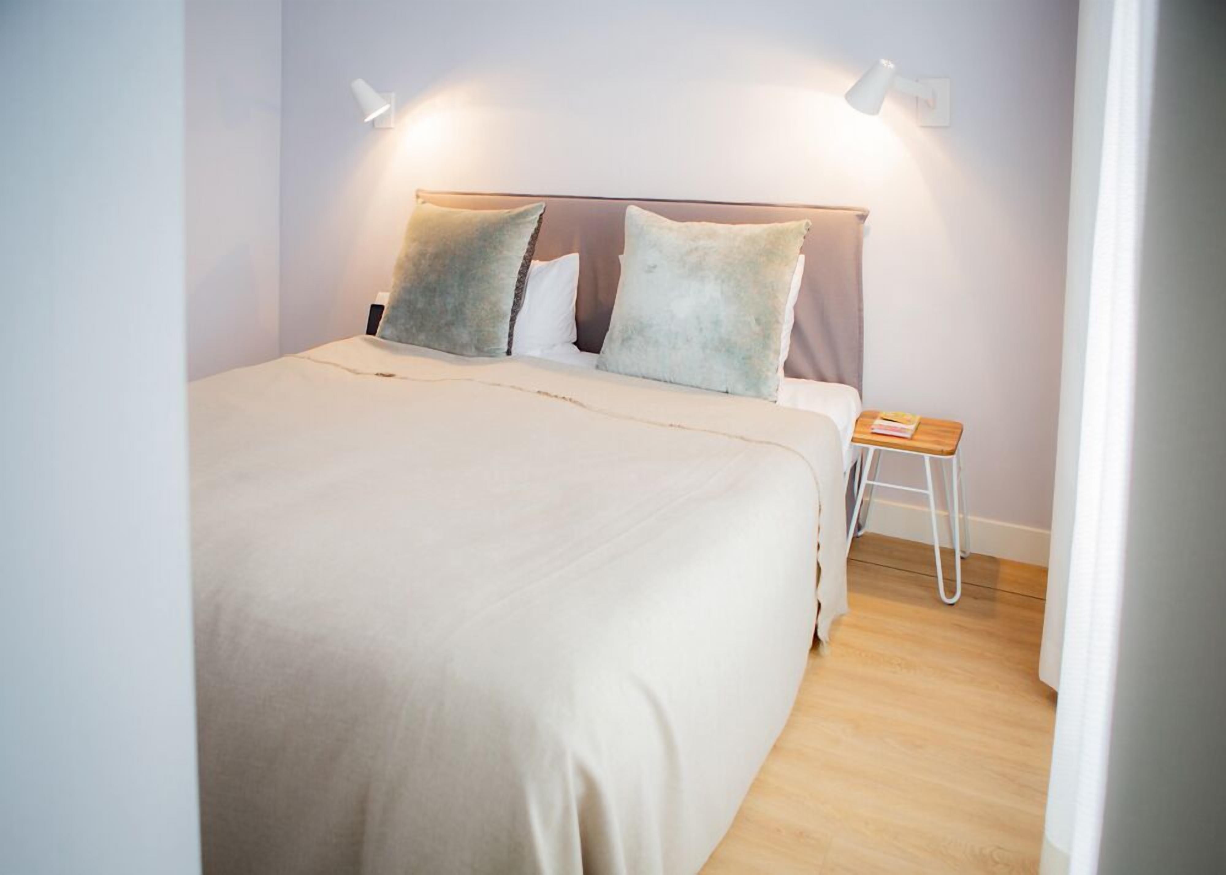 A neatly made bed with a white comforter and two pillows, a small wooden stool, and two wall-mounted lamps.
