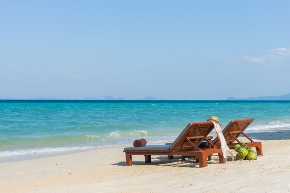 Dos sillones de madera sobre una playa arenosa con mar azul transparente y montañas al fondo.