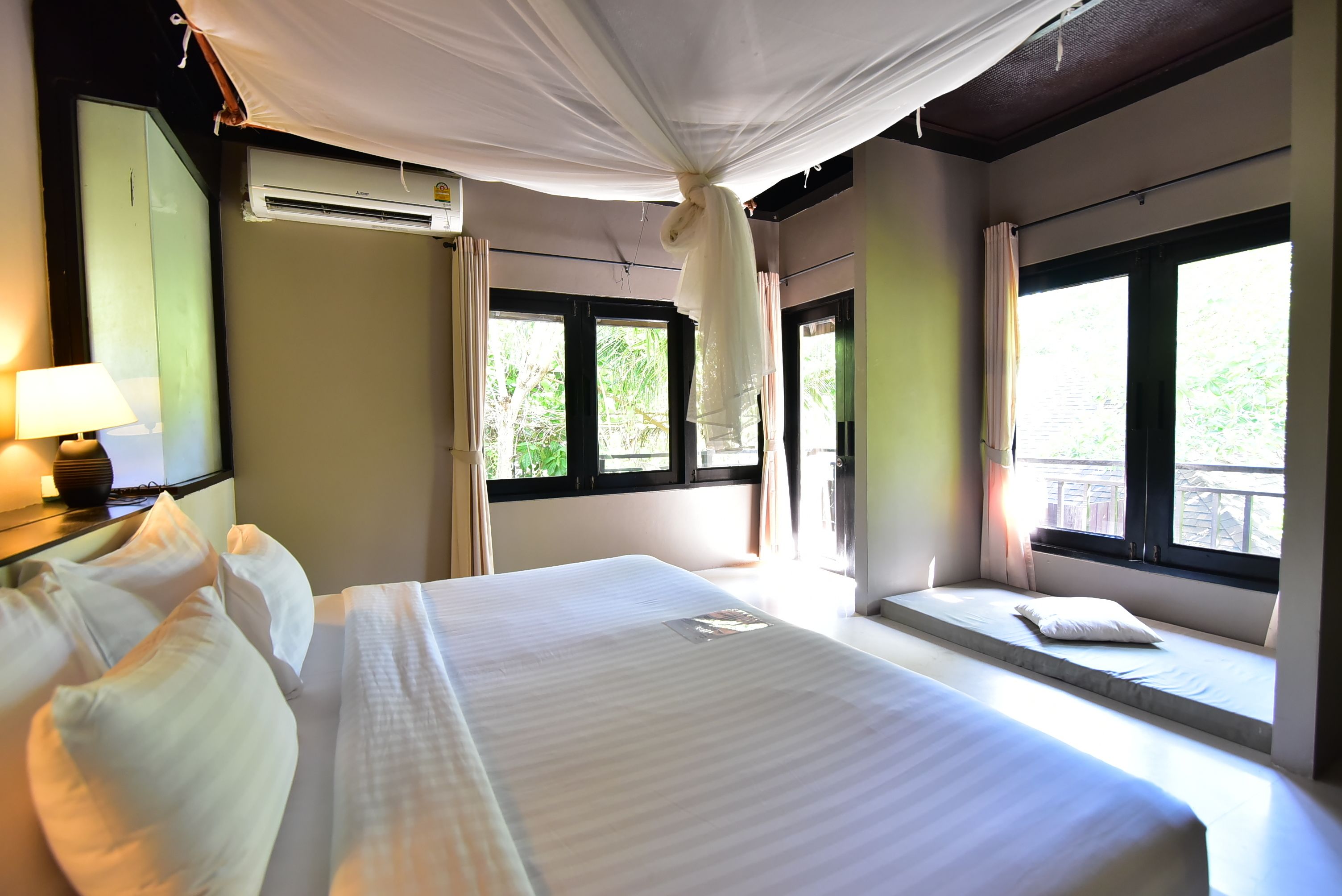 Una habitación de hotel con una cama grande, una mesita de noche con lámpara, una ventana con cortinas y un equipo de aire acondicionado.