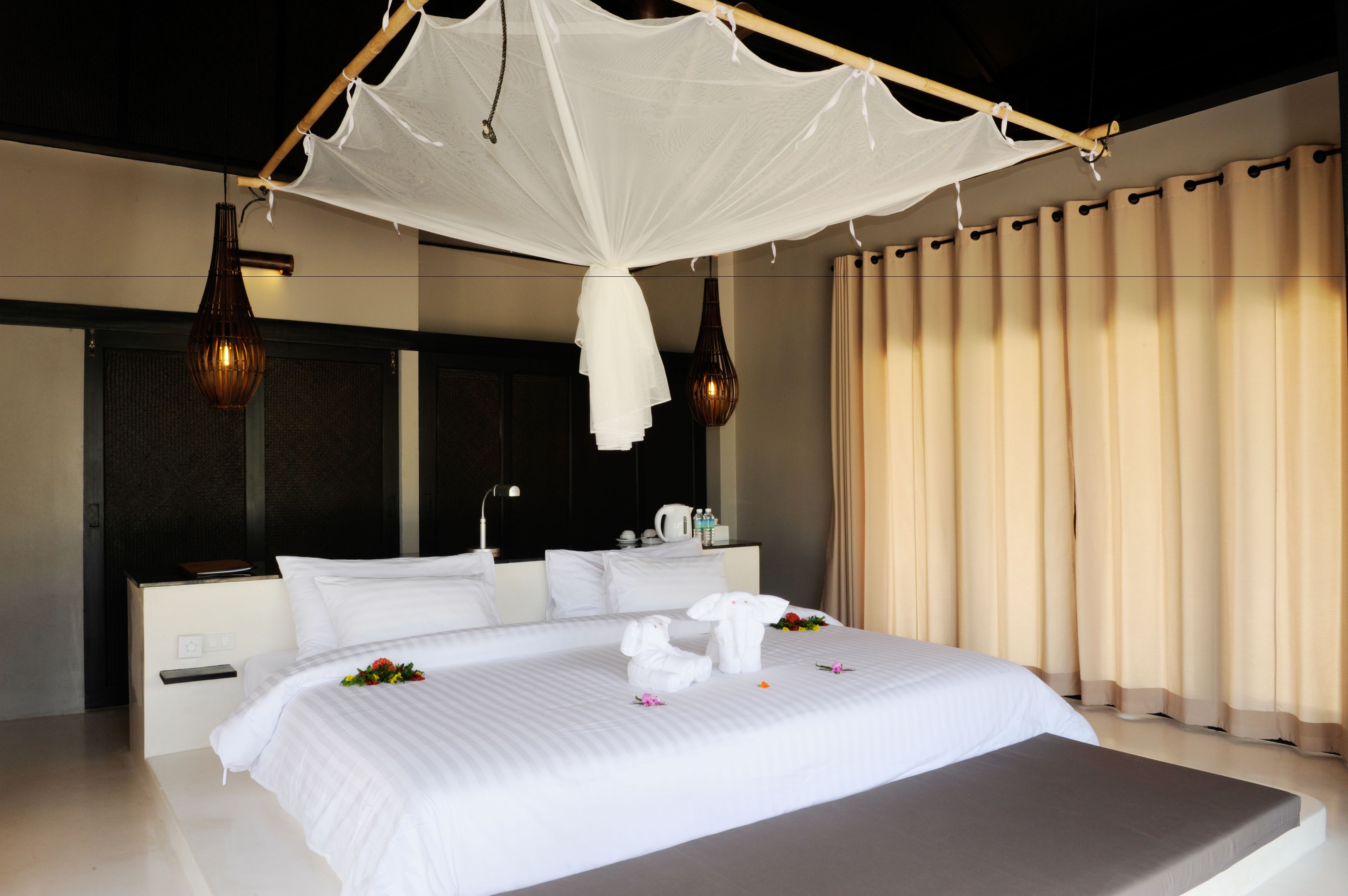 A neatly made bed with white linens and a canopy, a bedside table with a lamp, and a large window with curtains.