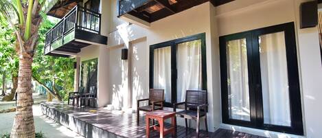 Una terraza de madera con sillas y una mesa, un balcón con barandilla y ventanas amplias con cortinas.