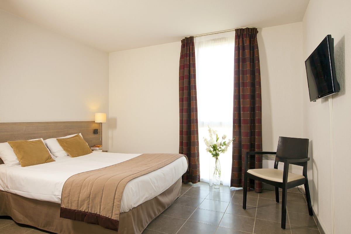 Una habitación de hotel con cama, una silla, un televisor y un ventanal con cortinas.