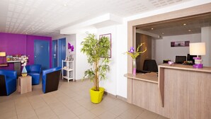 Een receptiezone met een houten toonbank, een computer en een stoel. Twee blauwe stoelen en een bijzettafeltje met een vaas staan er. In het midden staat een grote gele plant.