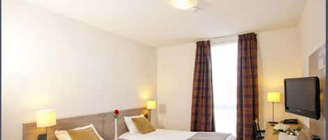 Habitación de hotel con dos camas, televisión, escritorio con lámpara y ventana con cortinas.