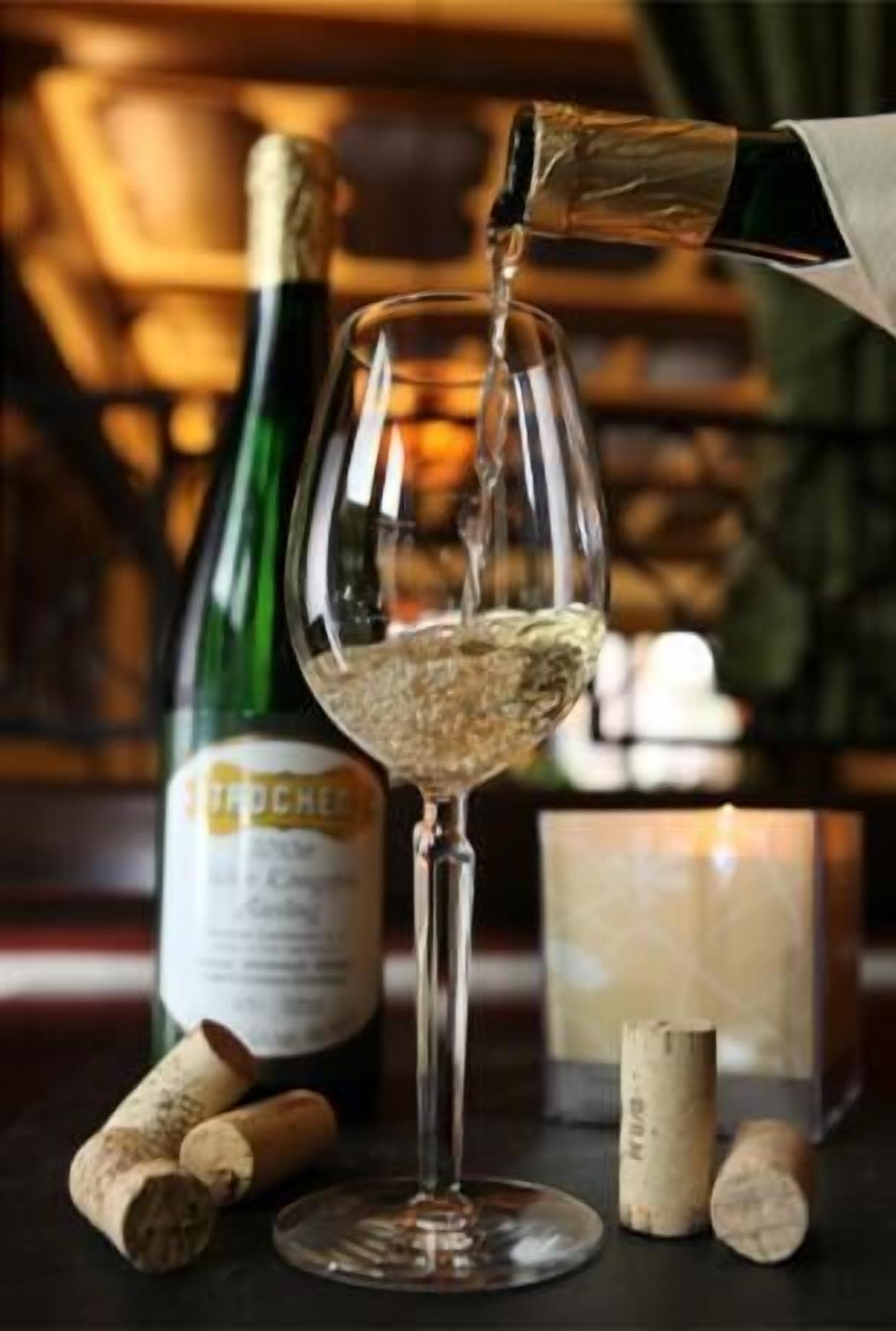 A wine glass being filled with a clear liquid, likely wine, from a bottle labeled "TRPCHEE". Corks are visible on the table, suggesting a wine tasting or serving setting.