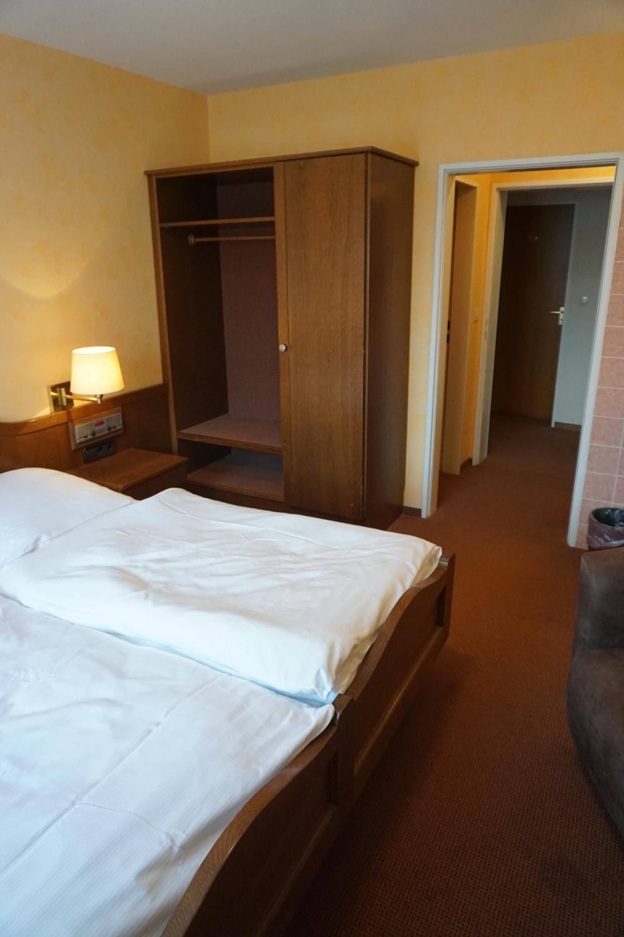A hotel room with a bed, a wooden wardrobe, a nightstand with a lamp, and a hallway leading to another room.